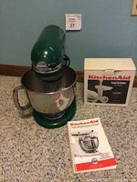 Green KitchenAid Artisan stand mixer with stainless steel bowl, KitchenAid food grinder box, and manual on beige carpet against teal wall.