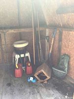 View of shed interior showing 2 red gas cans, wood bird feeder, green bird bath, assorted garden tools with wooden handles, and plastic tables.