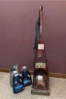 Full view of carpet cleaner with handle, power cord, and cleaner base unit alongside two jugs of cleaning solution on carpeted floor against wall.