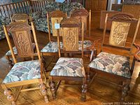 Photo showing a group of six wooden dining chairs including one captain chair, featuring carved backs with floral motifs and tapestry seats arranged on a hardwood floor.