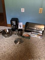 Overview of all small appliances and kitchen tools: FoodSaver vacuum sealer, roll of vacuum bags, white box of vacuum bag rolls, Sunbeam coffee maker, metal coffee filter mug, stainless steel food mill with masher attachments on carpet