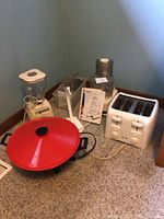 Photo of white Oster blender next to Cuisinart food processor parts and white 4-slice toaster alongside a red-lidded electric wok and banana holder