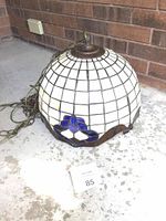 Full view of the dome-shaped Tiffany style stained glass lamp shade with electric cord around it placed on concrete floor near brick wall.