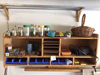 Wall-mounted wooden storage shelf with multiple compartments and blue plastic bins containing assorted nails, screws, bolts, staples, and wall anchors. Also visible are glass jars with fasteners, a woven basket, and tools including a Black & Decker planer.