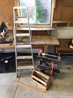 Photo of metal ladder, Workmate portable work bench, level, and wooden tool box arranged on a wooden bench and ground