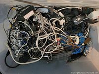 Overhead photo showing a large plastic bin filled with a tangled assortment of various cords and cables including USB, power adapters, Ethernet cables, speaker wire, computer mouse, and other electronic accessories.