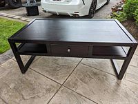 Full front view of the black rectangular coffee table showing flat top, drawer with knob, and side shelves.