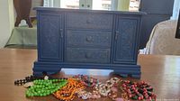 Front view of blue wooden jewelry box showing three central drawers and side cabinets, sitting on a table with some costume necklaces placed in front.