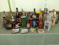 Full display of diverse empty bottles arranged in rows on green fabric, showing height and label differences. Front row includes decorative coasters.