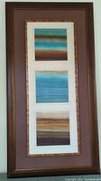 Framed wall art displayed vertically showing three rectangular abstract panels with blue and brown horizontal stripes.
