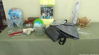 Wide photo showing an assortment of household items on a green cloth; includes glassware, storage containers, bags, kitchen gadgets, and rolled microfiber towels to the back.