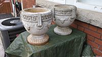 Two vintage cement planters placed on a green tarp outside against a brick wall. Front view showing overall shape, design, and weathered condition.