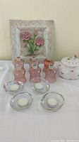 Photo showing six rose-colored glass candle holders, four clear glass candle holders with white tea lights, ceramic potpourri container with rose pattern, and rose-themed wall hanging.