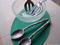 Pressed glass salad bowl on green mat with two long metal twig-style salad servers with leaf-shaped ends in front