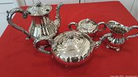 Four-piece silver plate coffee and tea set shown on a red background. Includes coffee pot, teapot, creamer, and sugar bowl with lids and handles. Reflective silver surfaces with decorative details.