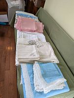 Photo of ten folded tablecloths and napkins on a green couch, showing various colors and lace details
