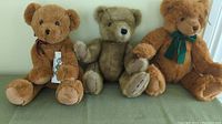 Photo of three teddy bears in a seated row on a green surface: one light brown with burgundy bow and tag, one medium brown with stitched paws, and one dark brown with green bow.