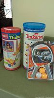 Two Tinkertoy sets in cylindrical containers and package of table tennis paddles and balls on green surface.