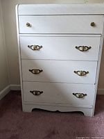 Photo of white wooden dresser with brass handles and knobs on four drawers, set on carpeted floor near wall corner.