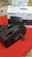 Front and side view of Pioneer wireless sound system placed on a red surface with original box and instructions behind it. Remote control is shown attached to the front of the system.
