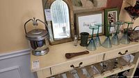 Top of white cabinet with stainless steel ice bucket, small arched brass mirror, small framed art, pitcher, green and clear stemmed glasses, and wooden bar tool