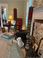 Pair of large brass candle holders and large red candles with patterned surface, small table lamp with beige shade on carpeted floor in living room.