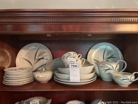 Full view of shelves with stacked plates, bowls, cups, and serving pieces showing plant motif and white glazed finish.