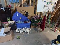 Wide view showing boxes, bins and bags of Christmas decorations including ornaments, garland, lights and artificial trees.