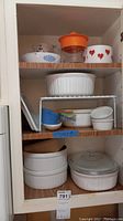 Several white ribbed Corning Ware casserole dishes on shelves, one with clear glass lid, plus smaller dishes including blue and white ceramic bowls and a red floral patterned bowl, glass pie plate with blue cornflower pattern, orange citrus juicer on top shelf.