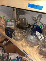 Shelf containing various clear and colored glass pitchers, bowls, and glasses, plus blue decorative glass item with rope handle.