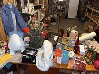 Wide view of assorted holiday decorations on a worktable including candleholders, figurines, plates, small artificial Christmas tree, picture frames, ribbons, mugs, and boxes with figurines.