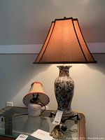 Full view of the pair of lamps on a glass table showing white ceramic lamp with floral design and beige shade, and a terra-cotta ceramic lamp with smaller beige and red trimmed shade.
