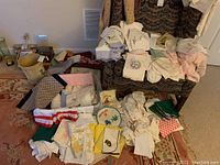 Wide view of assorted linens, embroidered cloths, patterned blankets and plastic picture frames on floor and couch.