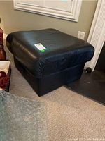 Black leather square ottoman shown from a side angle on carpet near window and other furniture, confirming size and condition.