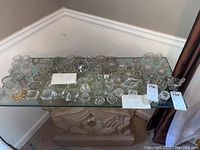 Over 50 pieces of clear cut glass and crystal salt cellars in a collection on a glass tabletop, showing various shapes including round, square, octagonal, some with pedestal bases, and one piece with a yellow tint.