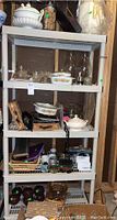 Five shelf white IKEA plastic shelving unit holding various servingware items including Corningware casserole dishes and soup tureen with Spice of Life pattern, glassware, baskets, and candleholders.