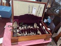 Wooden chest containing assorted silverplate flatware pieces arranged in rows with individual slots and loose pieces, burgundy velvet lining visible, with knives in fabric lined upper section.