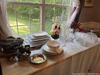 Wide view of assorted silver plated serveware, ceramic plates, glassware and decorative items displayed on white tablecloth by window with outdoor view
