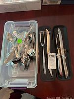Full view showing the container with assorted stainless steel flatware including spoons, forks, knives, and the black case containing the carving set with knife, fork and sharpening steel.