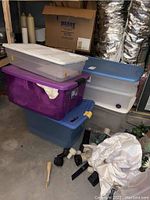 Stack of six large plastic storage bins of various colors with lids in a basement setting, visible surrounding area shows furnace ducts and other items.