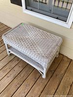White wicker ottoman placed on wooden porch flooring with woven wicker surface and shelf below.