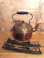Photo of a hammered copper kettle with a large curved spout and black handle, placed on a wooden surface. In front of the kettle is a pair of antique metal speed skates with adjustable straps, showing wear and age.