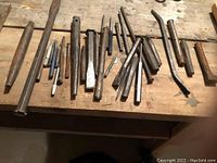 Photo of assorted metal punches and chisels on wooden workbench, showing various sizes and shapes including flat chisels, pointed punches and bent chisels, all in used condition.