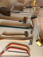 Photo of assorted tools laid out on a surface, includes shovels, axes, hack saws, a pick axe, a weed trimmer handle, and a digging bar.