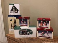 Photo of three boxed Lemax Christmas village accessories including a tall Christmas tree, a horse-drawn dairy sleigh, and a snowball fight scene.