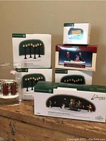 Photo showing boxed Department 56 Christmas village accessories including park bench, street lanterns, sleighs, and Cratchit family caroling figurines.
