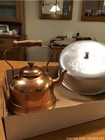Photo showing a copper kettle with a wooden handle next to a silver tin cake lid with embossed floral pattern and top handle.