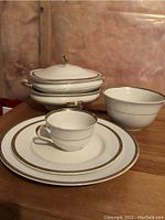 Photo shows stack of two oval serving dishes, one with lid, along with one bowl, dinner plate, luncheon plate, and cup with saucer.