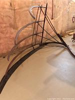 Large antique cradle scythe resting on a floor with insulating material background. Shows wooden curved arms, metal catcher rods and blades.