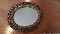 Photo of a round mirror placed on a wooden floor, showing its ornate gold/brass coloured carved frame and clear mirror glass.